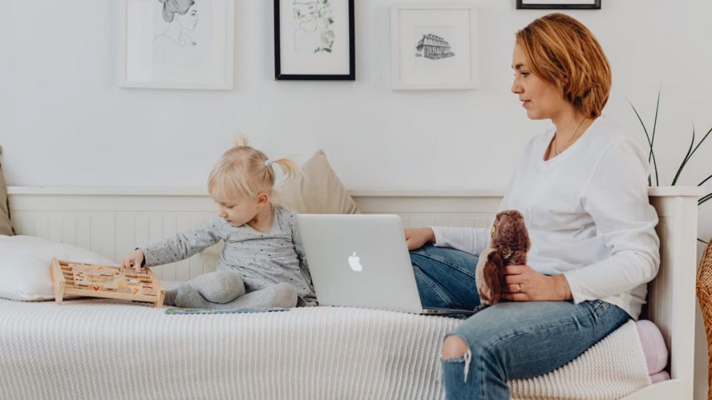 A mother and daughter bonding indoors, sitting on a sofa with a laptop and wooden toy.